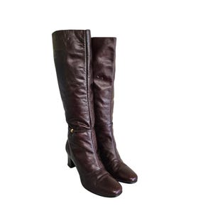 Puede incluir: Un par de botas altas de cuero marrón. Las botas tienen punta cuadrada, tacón pequeño y un detalle dorado alrededor del tobillo. El cuero parece ligeramente desgastado, dándoles un aspecto vintage. Las botas están de pie.