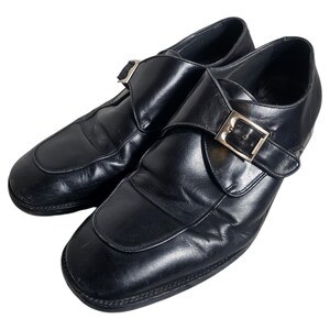 Pode incluir: Um par de sapatos de monge de couro preto com fivela dourada. Os sapatos t&ecirc;m bico fino e salto baixo.