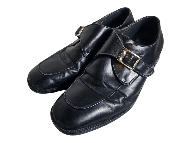 Pode incluir: Um par de sapatos de monge de couro preto com fivela dourada. Os sapatos t&ecirc;m bico fino e salto baixo.