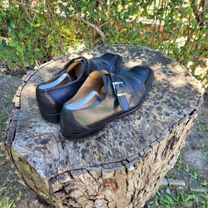 K&ouml;nnte beinhalten: Ein Paar schwarze Lederschuhe mit Schnallenverschluss. Die Schuhe stehen auf einem Baumstumpf.