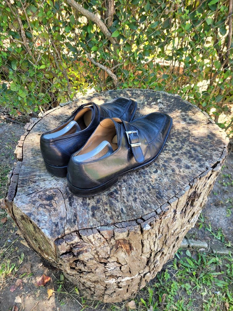 K&ouml;nnte beinhalten: Ein Paar schwarze Lederschuhe mit Schnallenverschluss. Die Schuhe stehen auf einem Baumstumpf.