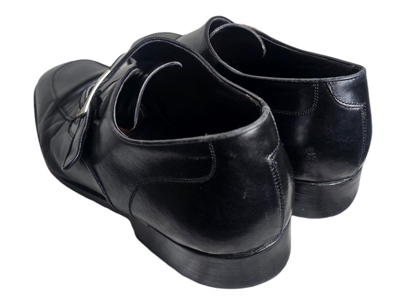 Pode incluir: Um par de sapatos de couro preto com fecho de fivela. Os sapatos t&ecirc;m bico fino e salto baixo.