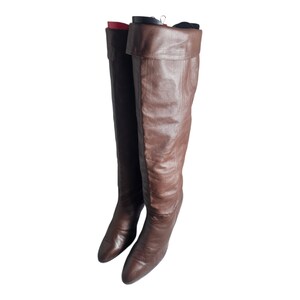 May include: A pair of brown leather knee-high boots with a pointed toe and a low heel. The boots are hanging on a hanger.