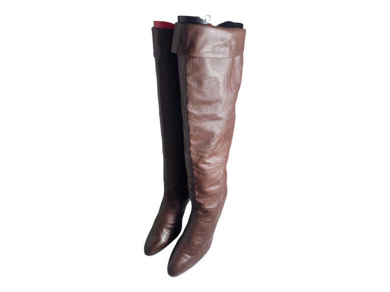 May include: A pair of brown leather knee-high boots with a pointed toe and a low heel. The boots are hanging on a hanger.