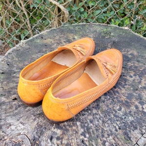 May include: A pair of brown leather loafers with tassels. The loafers have a woven pattern and are sitting on a wooden surface.