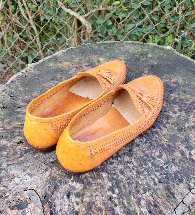 May include: A pair of brown leather loafers with tassels. The loafers have a woven pattern and are sitting on a wooden surface.