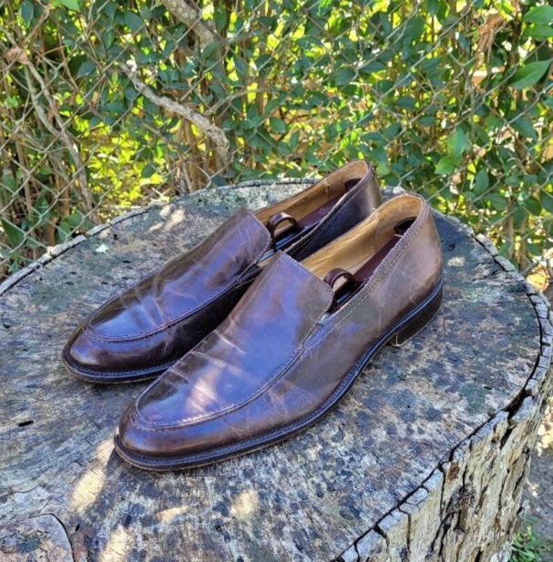 Pode incluir: Um par de sapatos marrons de couro sem cadar&ccedil;os com um visual ligeiramente desgastado. Os sapatos t&ecirc;m uma ponta pontiaguda e um salto baixo.