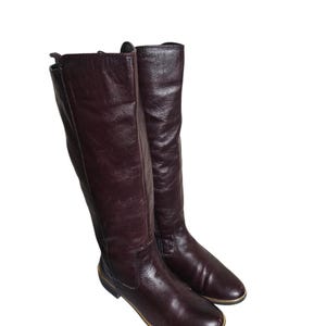 May include: A pair of knee-high boots crafted from dark burgundy leather. These boots feature a smooth, rounded toe, a flat sole with a thin gold-toned trim along the base, and a vertical side zipper closure extending from the ankle to the top.