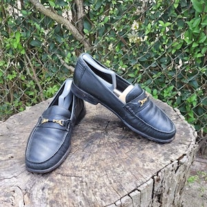 May include: A pair of black leather loafers with gold chain accents. The loafers are sitting on a tree stump.