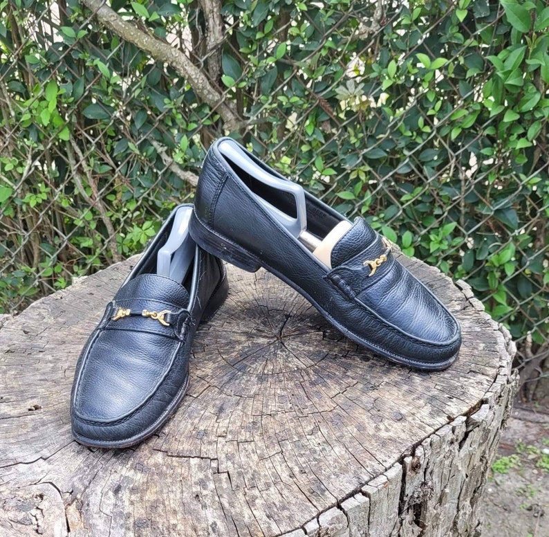 May include: A pair of black leather loafers with gold chain accents. The loafers are sitting on a tree stump.