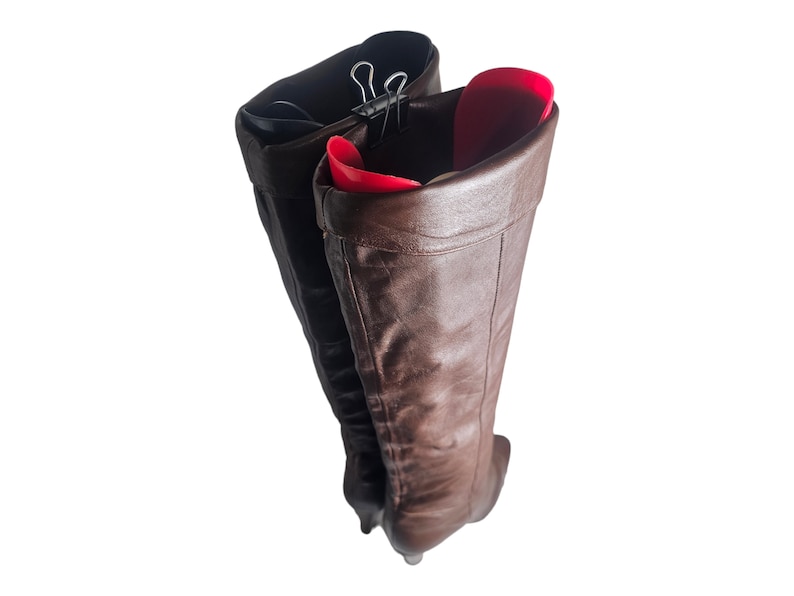 May include: A pair of brown leather boots with a pointed toe, held together with a metal clip. The boots are folded down at the top, revealing a red lining.