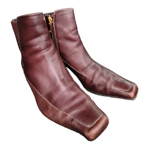 May include: A pair of brown leather ankle boots with square toes. The boots have a side zipper with a gold-colored zipper pull and white stitching details. The boots are set against a white background.