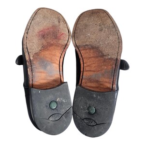 Pode incluir: Um par de sapatos de couro preto com solas de couro marrom. Os sapatos t&ecirc;m um fecho de fivela e um salto de borracha.
