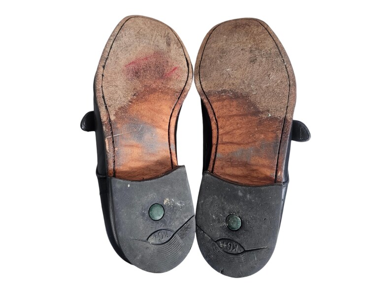 Pode incluir: Um par de sapatos de couro preto com solas de couro marrom. Os sapatos t&ecirc;m um fecho de fivela e um salto de borracha.