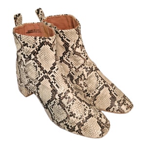 May include: A pair of beige ankle boots with a snakeskin pattern. The boots have a block heel and a pointed toe. The label on the inside of the boot reads "Made in Chile / Fabrique en Chile."