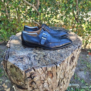 K&ouml;nnte beinhalten: Ein Paar schwarze Monk-Strap-Schuhe aus Leder mit einer silbernen Schnalle. Die Schuhe stehen auf einem Baumstumpf.