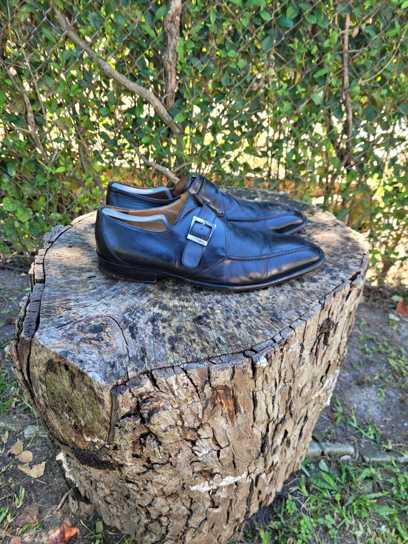 K&ouml;nnte beinhalten: Ein Paar schwarze Monk-Strap-Schuhe aus Leder mit einer silbernen Schnalle. Die Schuhe stehen auf einem Baumstumpf.