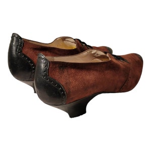 May include: A pair of brown suede heeled shoes with black leather trim and laces. The shoes have a low heel and a rounded toe.