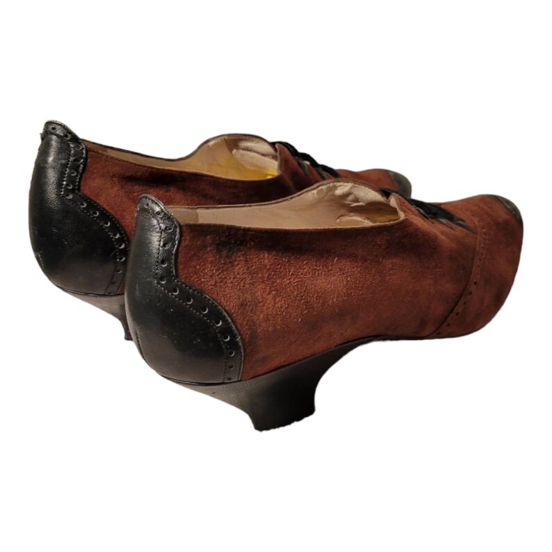 May include: A pair of brown suede heeled shoes with black leather trim and laces. The shoes have a low heel and a rounded toe.