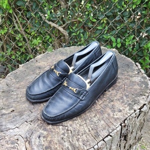 May include: A pair of black leather loafers with gold buckles. The loafers are on a wooden surface.