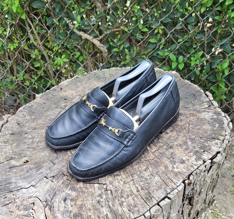 May include: A pair of black leather loafers with gold buckles. The loafers are on a wooden surface.