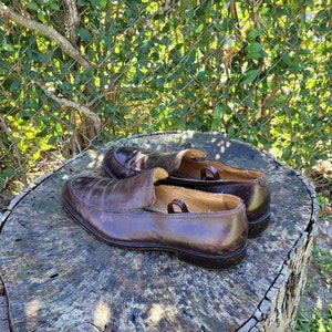 Pode incluir: Um par de sapatos de couro marrom em um tronco de &aacute;rvore. Os sapatos t&ecirc;m um design de deslizamento e uma ponta arredondada.