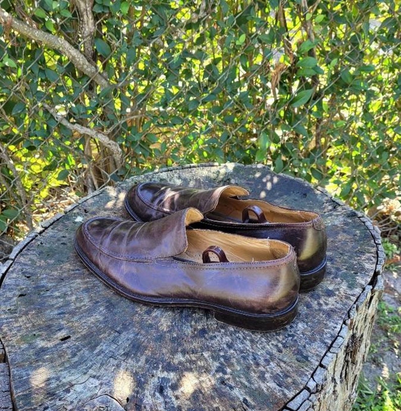 Pode incluir: Um par de sapatos de couro marrom em um tronco de &aacute;rvore. Os sapatos t&ecirc;m um design de deslizamento e uma ponta arredondada.