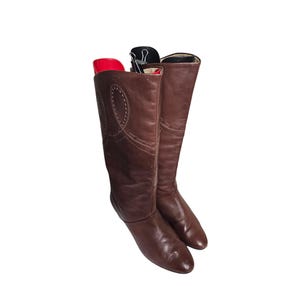 Puede incluir: Un par de botas altas de cuero marrón. Las botas tienen la punta afilada y un diseño decorativo cosido en el lateral. Las botas están vacías y tienen hormas rojas y negras.