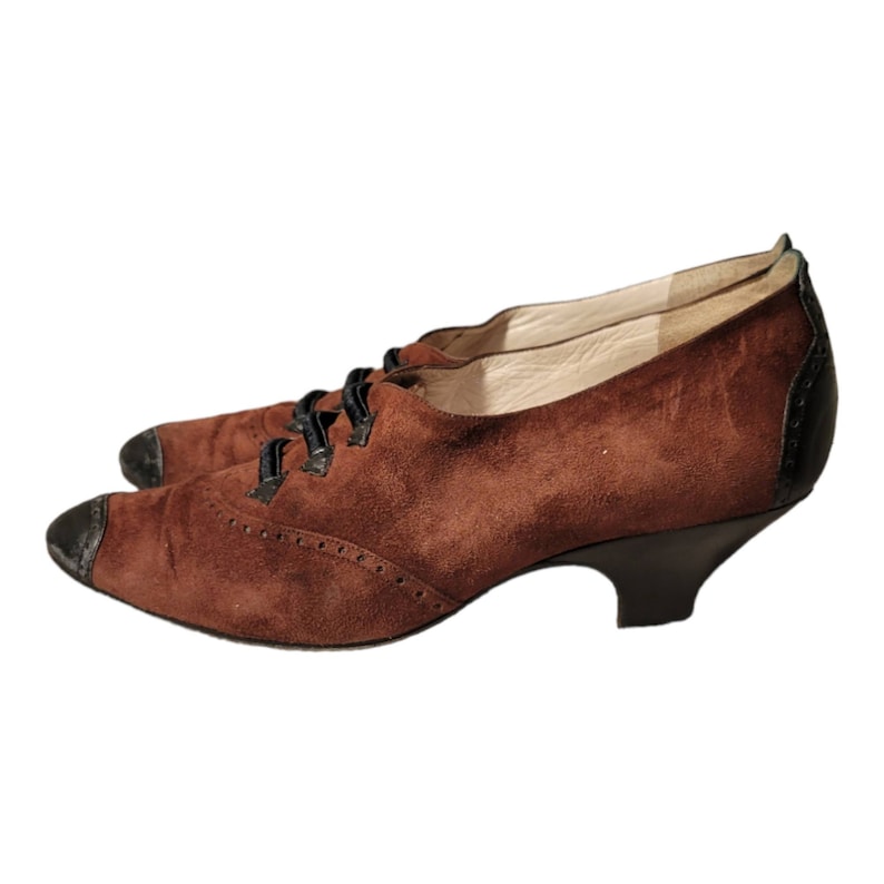 May include: Brown suede heeled shoes with black laces and a black heel. The shoes have a brogue design.