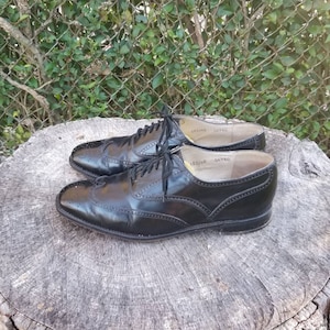 Puede incluir: Un par de zapatos negros de cuero con cordones y detalles de punta de ala. Los zapatos tienen un acabado pulido y est&aacute;n sobre un toc&oacute;n de &aacute;rbol.