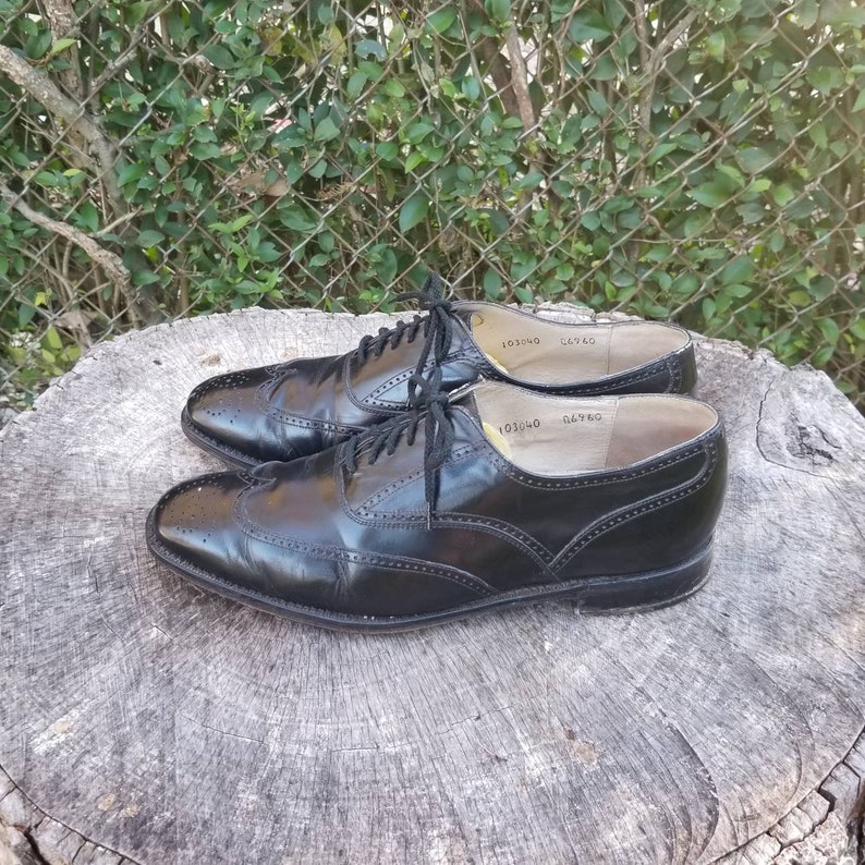 Puede incluir: Un par de zapatos negros de cuero con cordones y detalles de punta de ala. Los zapatos tienen un acabado pulido y est&aacute;n sobre un toc&oacute;n de &aacute;rbol.
