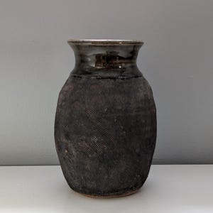 May include: A dark brown ceramic vase with a textured surface. The vase has a wide, rounded body and a narrower neck with a dark, glossy rim. The textured surface has a pattern of vertical lines.