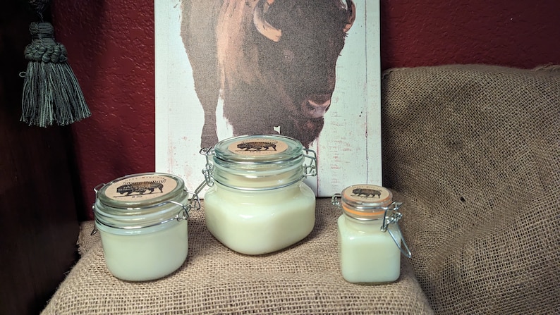 May include: Three glass jars with lids containing white candles. The jars have a brown label with a black silhouette of a buffalo on them. The jars are sitting on a burlap surface in front of a painting of a buffalo.