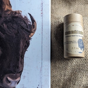Può includere: Un tubo di cartone marrone con un'etichetta bianca che recita "Bison Solid Balm" e "Moisture Seal For Dry Skin". L'etichetta presenta anche una piccola immagine di un bisonte. Il tubo è appoggiato su una superficie di juta marrone.