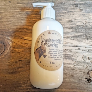 Può includere: Una bottiglia bianca con pompa di crema di latte d'asina con un'etichetta marrone che presenta un'illustrazione di un asino e il testo "Soapy Aks Donkey Milk Cream". L'etichetta elenca anche gli ingredienti e indica "8 oz. Made in Texas".