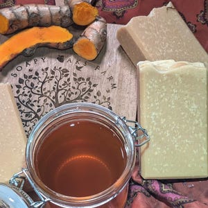 May include: Three bars of handmade soap with a light brown color and a grainy texture. The soap is on a wooden surface with the text "Soapy Acres Farm" printed on it. There are pieces of turmeric root and a jar of honey in the image.