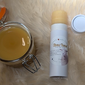 May include: A white tube of "Elbow Rescue" Rough Skin Treatment by Soapy Oaks Farm. The tube is labeled with ingredients including: Natural Lemon, Shea Butter, Coconut Oil, Jojoba Oil, Vitamin E, and Lavender Essential Oil. The tube is 3 oz and made in Texas. A clear glass jar with a yellow liquid and an orange lid is also in the image.
