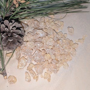 May include: A pile of translucent, light amber-colored resin pieces, resembling small, irregular crystals. The resin is scattered on a light beige surface, with a pine branch and pine cone in the background.