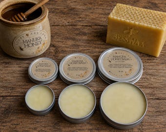 May include: A collection of skincare products including a ceramic jar of Manuka honey with a wooden dipper, a rectangular block of beeswax, and three round metal tins of Manuka Honey Balm in 1 oz, 2 oz, and 4 oz sizes with visible labels.