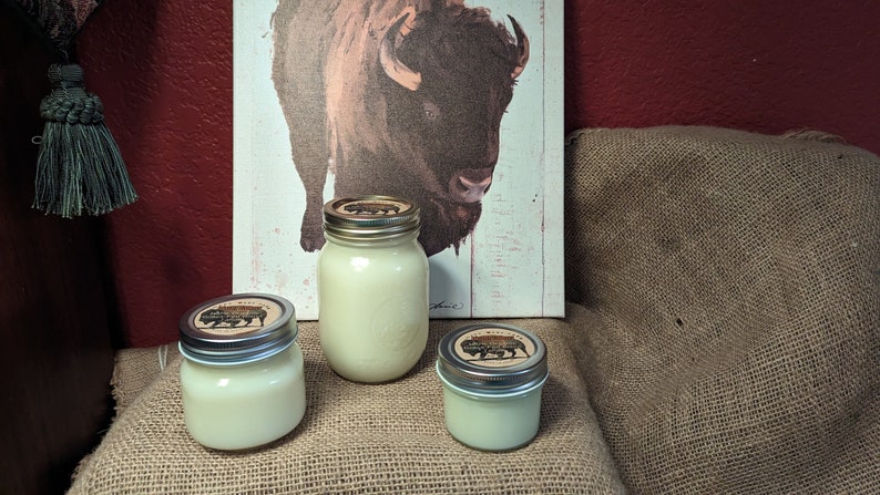 May include: Three glass jars with white candles and brown lids. The lids have a black and white image of a buffalo on them. The jars are sitting on a brown burlap surface.