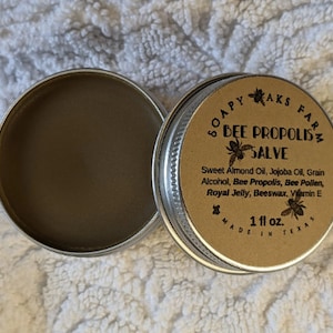 May include: A small, round, metal tin with a brown label that reads "SOAPY AKS FARM BEE PROPOLIS'z SALVE". The label lists the ingredients: Sweet Almond Oil, Jojoba Oil, Grain Alcohol, Bee Propolis, Bee Pollen, Royal Jelly, Beeswax, Vitamin E. The label also states "1 fl oz. MADE IN TEXAS".