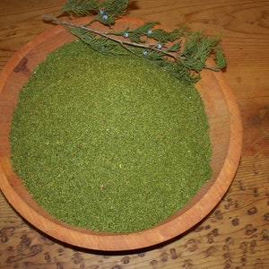 May include: A wooden bowl filled with dried green juniper berries. A sprig of juniper with blue berries is resting on the rim of the bowl.