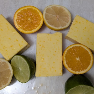 May include: Five bars of yellow soap with a citrus scent, next to a lemon, two limes, and two oranges.