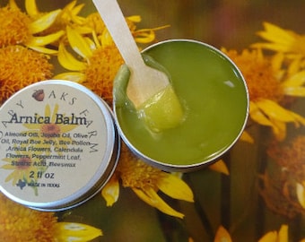 Puede incluir: Una pequeña lata plateada con tapa verde que contiene bálsamo de árnica. La etiqueta dice "Soapy AKS Farm Arnica Balm" y enumera los ingredientes: aceite de almendras, aceite de jojoba, aceite de oliva, jalea real de abeja, polen de abeja, flores de árnica, flores de caléndula, hoja de menta, ácido esteárico, cera de abejas. La lata está sobre una superficie cubierta de flores amarillas.