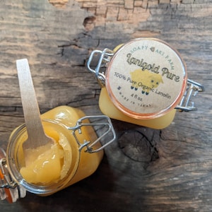 Könnte beinhalten: Zwei Glasbehälter gefüllt mit einer gelben Substanz, beschriftet mit "Lanigold Pure 100% Reine Bio-Lanolin 4 fl oz Hergestellt in Texas". Ein Holzlöffel befindet sich in einem der Behälter.
