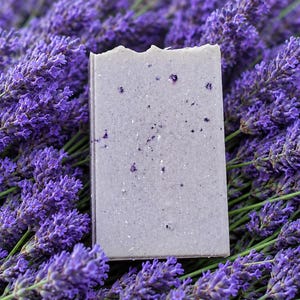 May include: A rectangular bar of light purple soap with a textured surface containing small dark purple botanical inclusions, positioned upright against a dense background of vibrant purple lavender flower stalks with green stems.