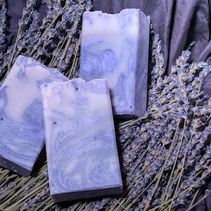 Può includere: Tre saponette alla lavanda con un designo marmorizzato viola e bianco. Il sapone è esposto su un letto di lavanda secca.