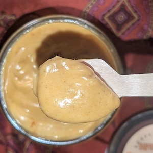 Puede incluir: Primer plano de una cuchara de madera sacando mantequilla de cacahuete espesa y de color marrón dorado de un tarro de vidrio. La mantequilla de cacahuete es suave y cremosa, con un ligero brillo.