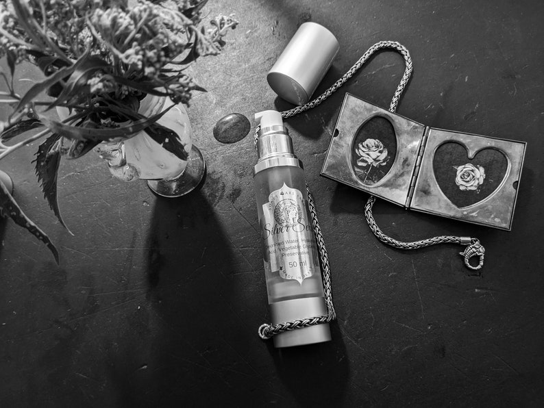 May include: A silver chain necklace with a silver locket featuring two heart-shaped compartments, each with a black and white image of a rose. A clear glass bottle with a silver pump top and a label that reads "Silver Serum" is on a dark surface. The bottle contains a clear liquid.