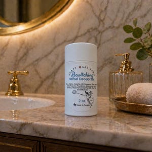 Peut inclure: Un contenant de déodorant cylindrique blanc avec le texte "Bewitching Herbal Deodorant" et une petite illustration. Le contenant est de 57 grammes et fabriqué au Texas. L'arrière-plan comprend un comptoir en marbre, des accessoires dorés et un plateau décoratif.